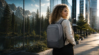 A woman with curly hair stands on a city sidewalk next to a reflective glass wall showing a mountain and forest scene. She is wearing a light gray backpack, which is the product being showcased in this lifestyle setting.