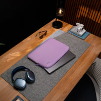 A lavender laptop sleeve with a matching zippered pouch rests on top of a closed silver laptop, placed on a gray felt desk mat on a wooden desk. A pair of blue wireless headphones, sunglasses, a black desk lamp, and a white instant camera are also visible in the scene.
