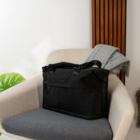 A black targus tote bag with top handles sits on a gray upholstered armchair in a modern interior setting. A folded gray blanket is draped over the back of the chair, and a wooden slatted wall panel is visible behind it.