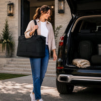 A woman carrying a large black duffel bag walks beside an open black SUV trunk in a driveway. The trunk contains a matching black backpack and a folded beige blanket.