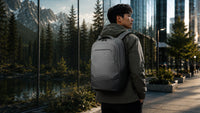 A man wearing a gray backpack stands on a city sidewalk in front of a large glass wall reflecting mountains and pine trees.