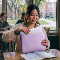 A woman in a beige knit sweater smiles while holding a lavender laptop sleeve with a white laptop inside, set in a bright cafe or study with a window showing a blurred campus background.