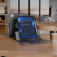 A tablet in a dark gray protective case with an attached keyboard and kickstand, shown on a wooden desk in a modern office setting. A coffee cup sits to the right, and the tablet screen displays a messaging app.