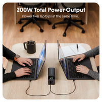 A portable black power station connected to two laptops on a light wood desk in an office setting, with text at the top reading '200W Total Power Output' and 'Power two laptops at the same time.'.